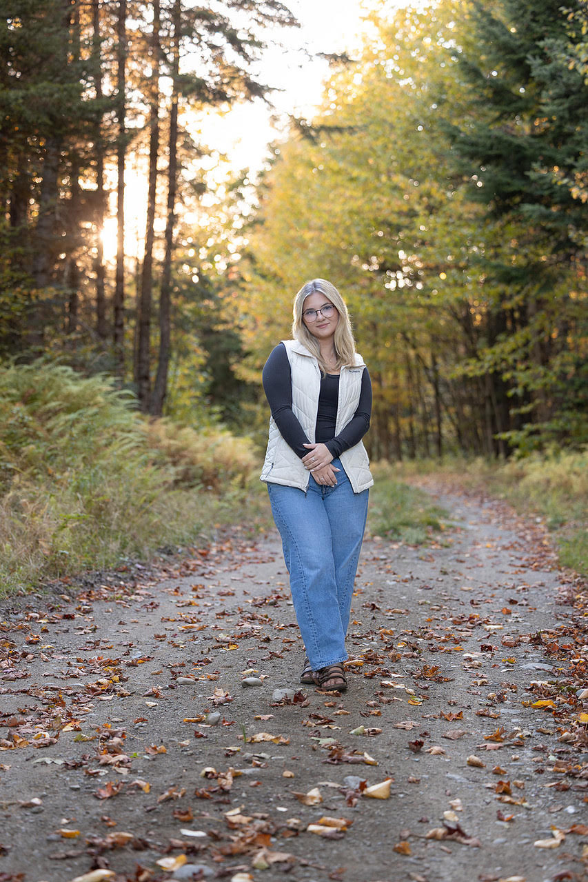 young woman in jeans, dark shirt, and white vest in paved path in a wooded area