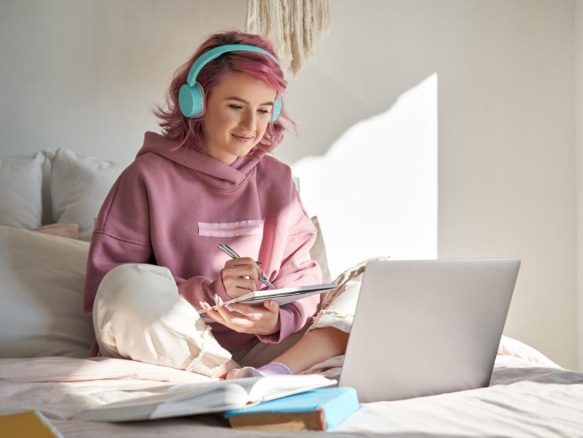 Young woman with headphones writing in notebook and looking at laptop